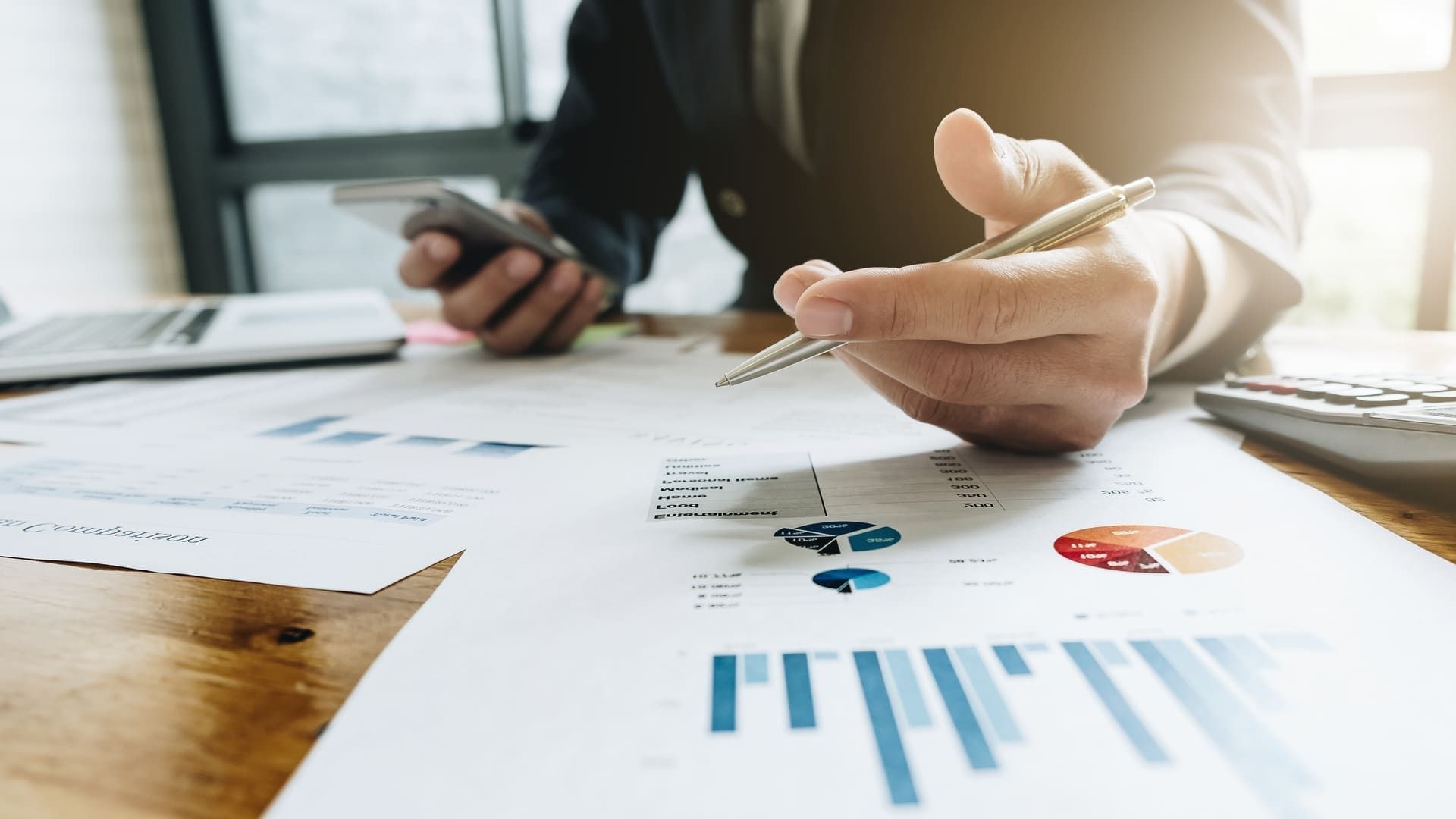 Finanzberater rechnet am Schreibtisch mit Taschenrechner in der Hand auf einem Stapel Dokumente mit graphischen Tabellen